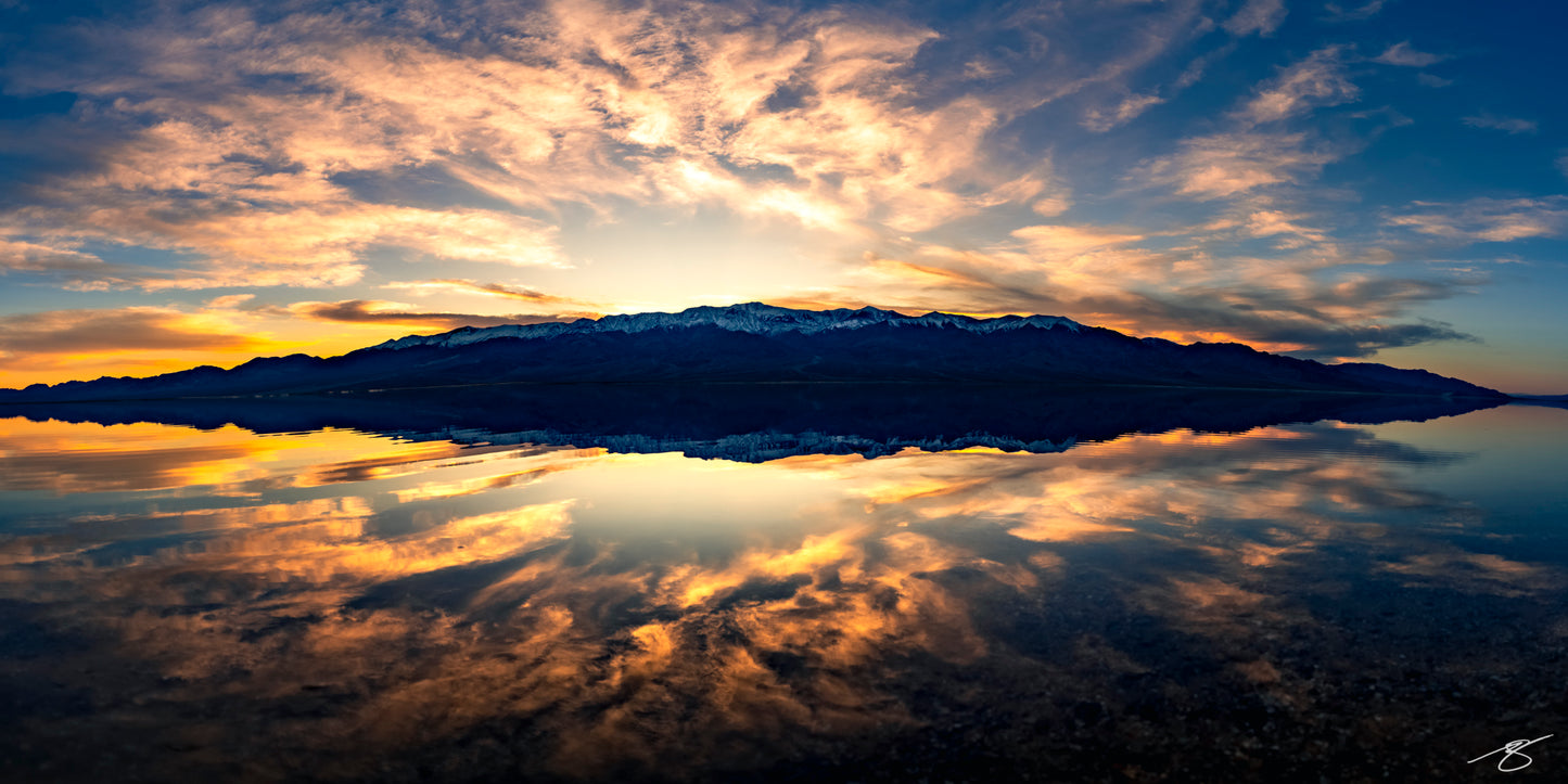 Wide panorama of sunset over a snow-capped mountain range reflected perfectly in a still lake, fiery clouds and deep blue sky creating a mirror-like scene