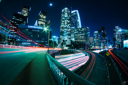 Panoramic night photo of downtown Los Angeles with skyscrapers glowing and red/white freeway light trails curving through the city; modern urban fine art print