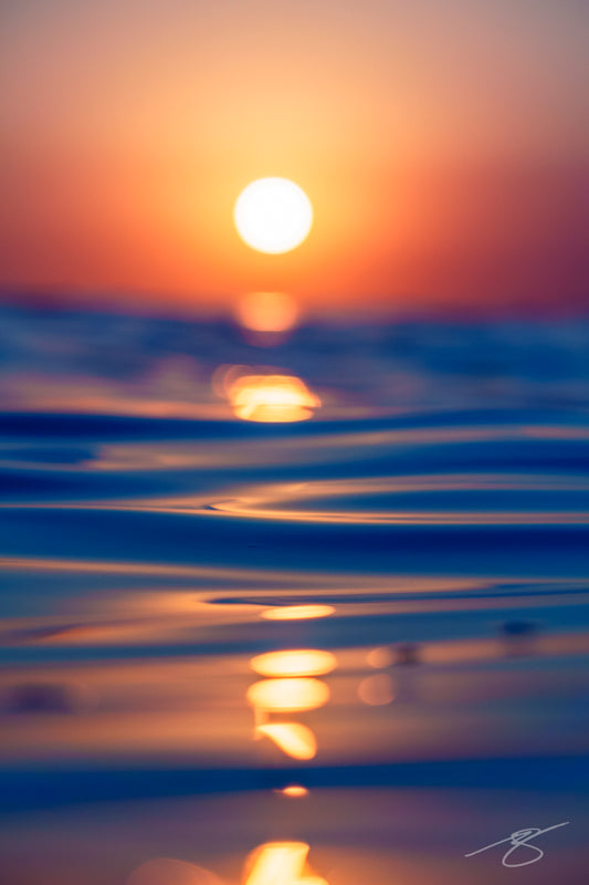 Waterline view of a glowing sunset with a golden reflection on gentle, rippled ocean water; minimalist seascape fine art photograph
