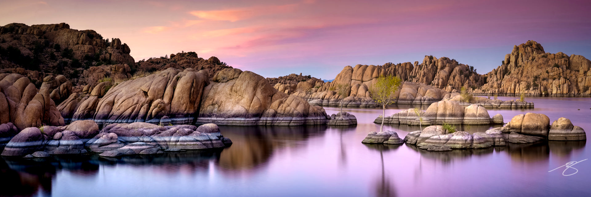 Panoramic long-exposure of Watson Lake’s Granite Dells at sunset, pastel pink-lavender sky reflecting on still water with rounded boulders and small trees