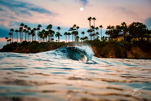 Waterline photo of a small barrel wave at dusk with a full moon above palm trees along a rocky cove; tranquil coastal fine art photograph