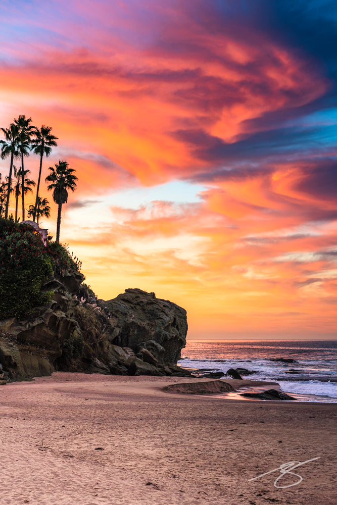 Sunset over a sandy cove with palm trees on a rocky bluff, pastel clouds filling the sky and gentle waves along the shore; coastal fine art photograph