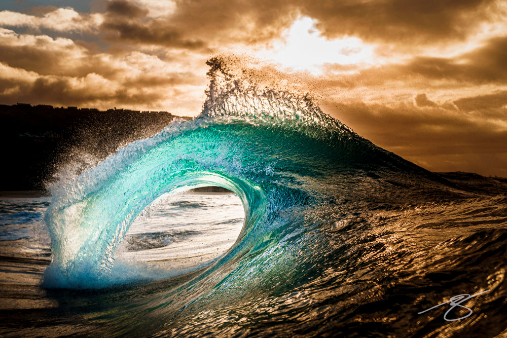 Backlit aqua barrel wave with golden spray at sunset beneath dramatic clouds; dynamic coastal fine art photograph