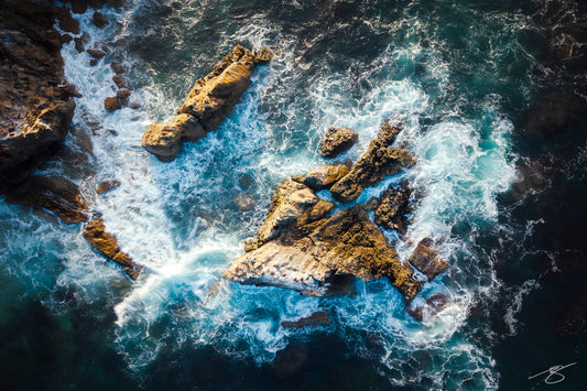 An overhead drone photograph of waves breaking against jagged coastal rock formations along the California shoreline. The image captures swirling turquoise water, foamy white surf, golden rock textures, and deep blue ocean shadows for a dramatic seascape.