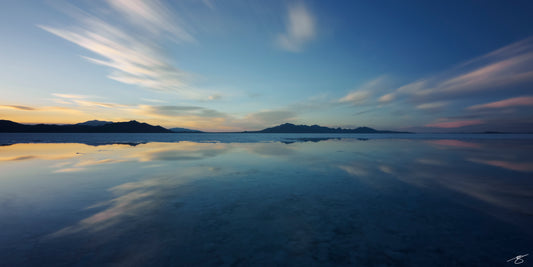 Peaceful long-exposure landscape of the Bonneville Salt Flats at sunset, featuring pastel clouds streaking across the sky, distant Utah mountains, and shallow reflective water creating a perfect mirrored horizon. Fine art desert photography.