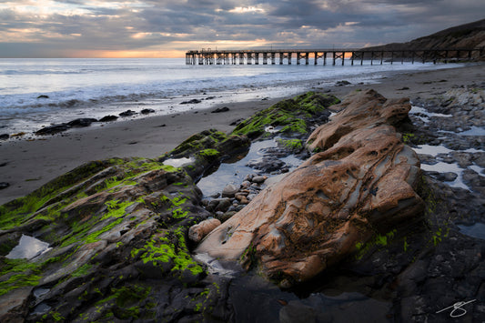 A coastal sunset scene in California featuring tide pools and moss-covered rock formations in the foreground. Soft waves roll toward the beach while a long wooden pier extends into the ocean under dramatic clouds, with warm evening light reflecting across the water.