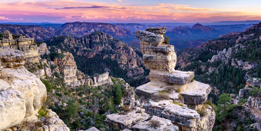 Panoramic view of a stacked rock hoodoo overlooking forested Arizona canyons at pastel sunset with distant mesas and pink clouds
