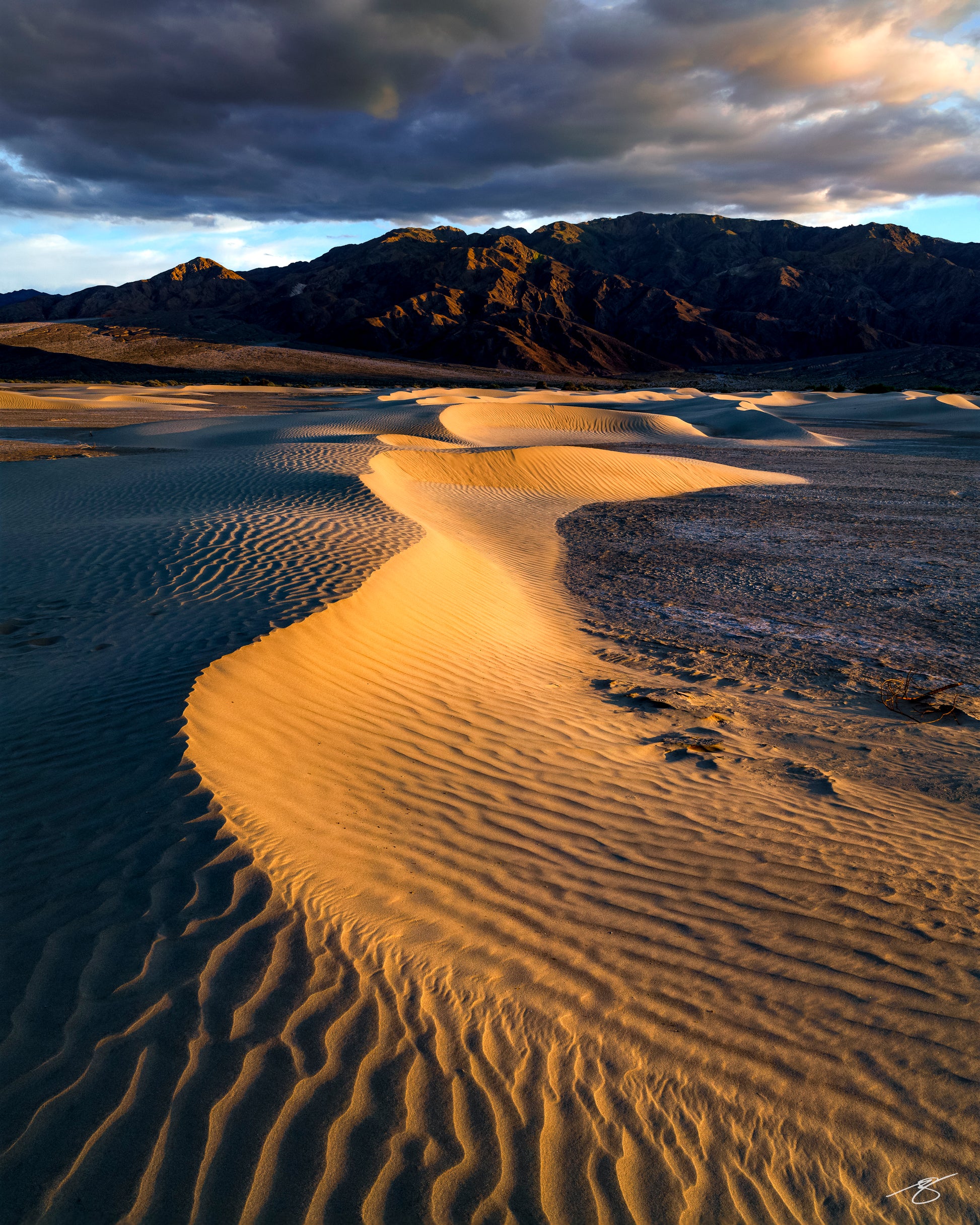 Fine art landscape of sunrise illuminating the sand dunes in Death Valley, featuring glowing golden ridges, sweeping wind-shaped textures, distant mountains, and dramatic clouds. A serene and elegant desert scene captured in warm morning light.