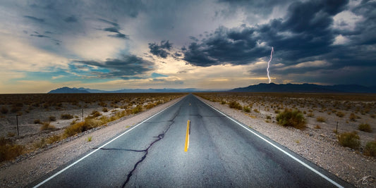 Straight desert highway leading to the horizon with storm clouds and a bright lightning strike over distant mountains, dramatic Southwest landscape