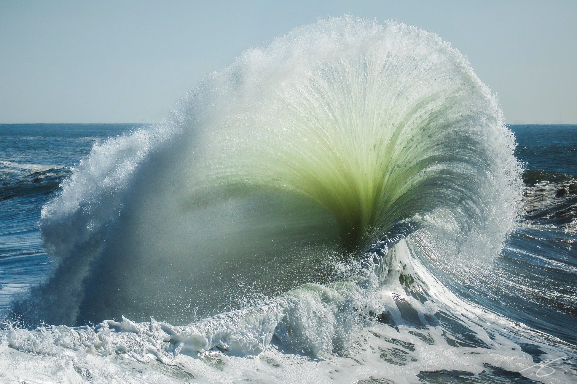 A dramatic photograph of an exploding wave captured at the peak of impact along the California coastline. A luminous green core radiates through the spray as thousands of water droplets burst upward, creating a sculpted fan-shaped explosion of ocean energy.