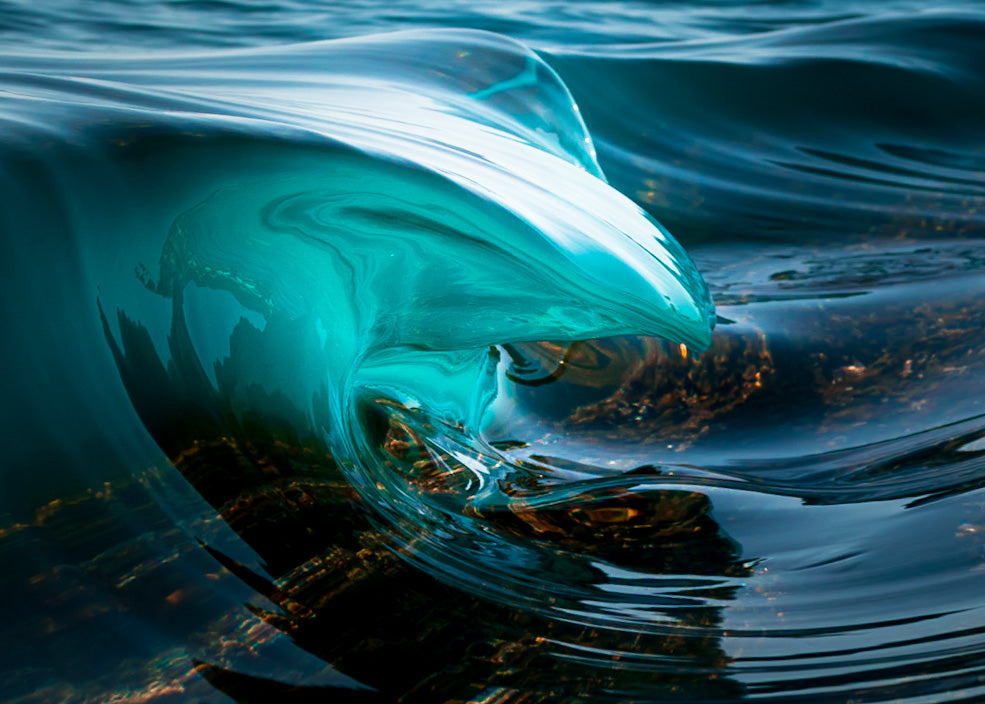 Close-up photo of a crystal-clear turquoise wave curling over a rocky reef, glassy surface and teal water creating an abstract coastal seascape