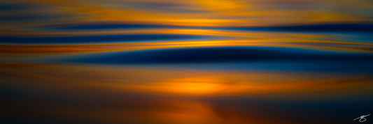 A minimalist abstract seascape showing deep blue ocean waves with warm golden reflections from the setting sun. The smooth, blurred motion creates layered horizontal bands of color, blending rich blue and molten gold to form a calming, modern coastal artwork.