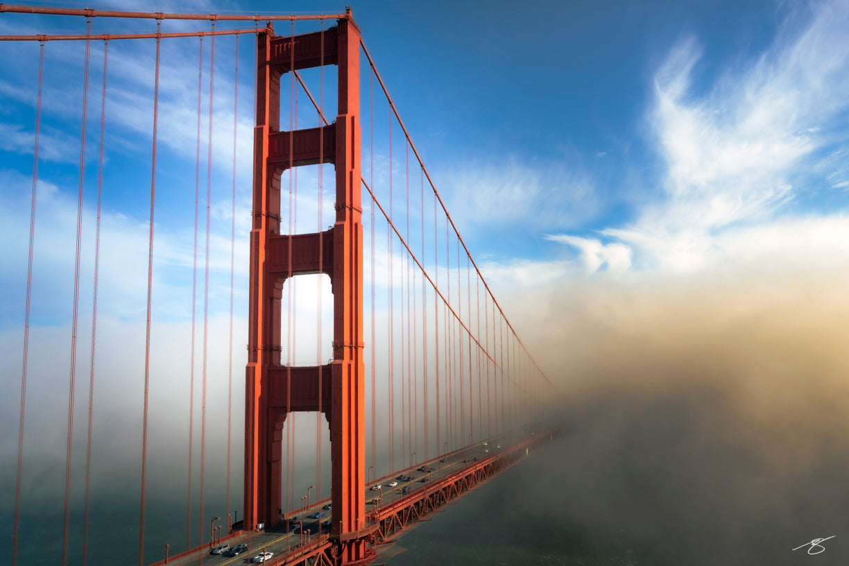 Fine art photograph of the Golden Gate Bridge rising above thick fog while its roadway disappears into the mist. The bright red tower contrasts with a blue sky and soft clouds, creating a dramatic and iconic San Francisco coastal scene.
