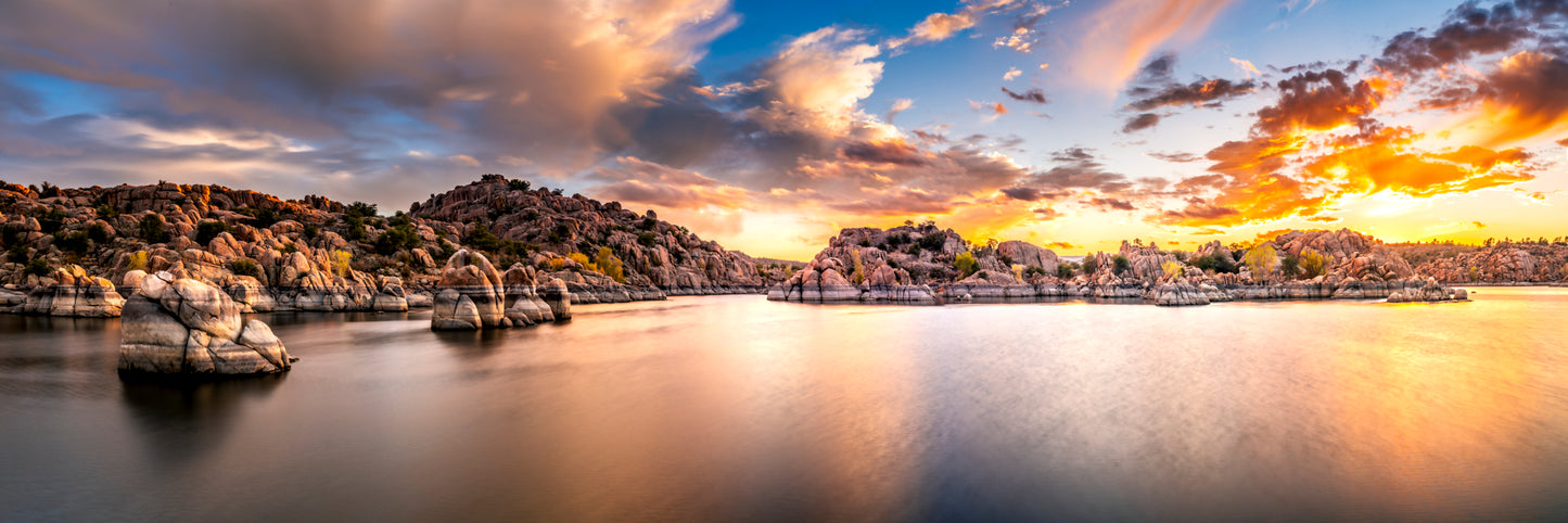 Panoramic sunset photograph of Watson Lake in Prescott, Arizona, showing smooth reflective water and the Granite Dells’ rounded pink boulders under glowing clouds