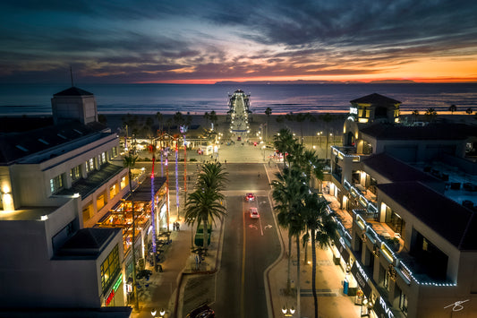 Fine art aerial photo of Huntington Beach at sunset, featuring the pier extending into the Pacific Ocean, glowing sky colors, illuminated downtown streets, palm trees, and warm coastal lighting. A vibrant and cinematic Southern California cityscape.