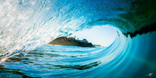 Inside a clear blue barrel wave looking toward a coastal bluff with palms and houses; immersive ocean fine art photograph
