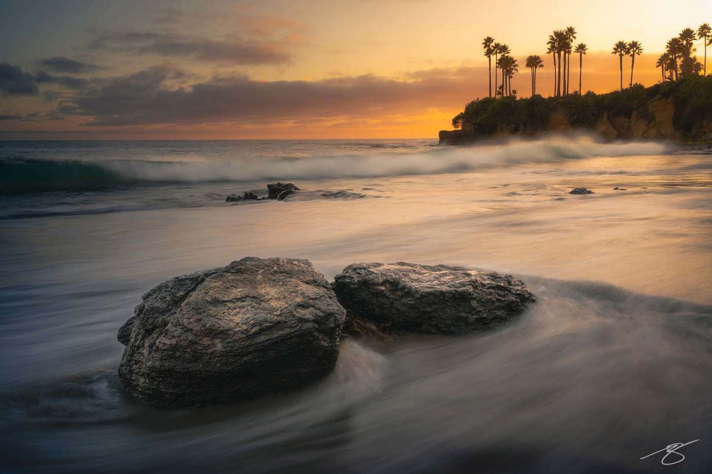 A peaceful sunset scene at Laguna Beach, California, featuring soft long-exposure waves moving around rocky shoreline foreground. Warm golden light illuminates the sky while tall palm trees stand silhouetted on the distant coastal cliffs, creating a serene seascape.