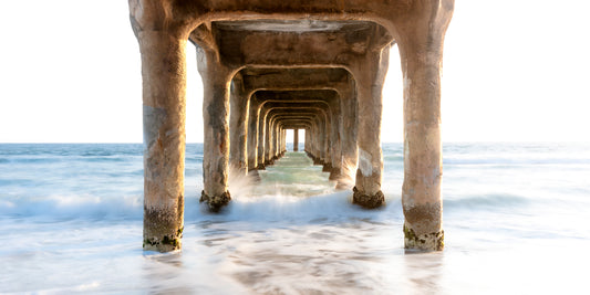 Long-exposure fine art photo of sunset beneath the Manhattan Beach Pier. Warm golden light glows through the concrete pillars while waves blur into soft motion around the structure, creating a serene and symmetrical California coastal scene.