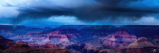Panoramic photo of a lightning strike beneath dark monsoon clouds over the Grand Canyon, revealing blue-purple layers of mesas and buttes at sunset