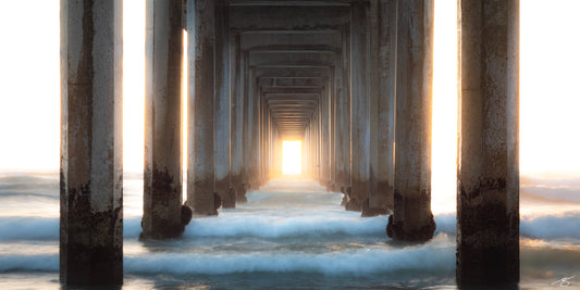 Long-exposure fine art photograph showing golden sunset light shining through Scripps Pier in La Jolla. Soft ocean waves move around the barnacle-covered pillars, creating a dreamy coastal scene with symmetry, motion, and warm California atmosphere.