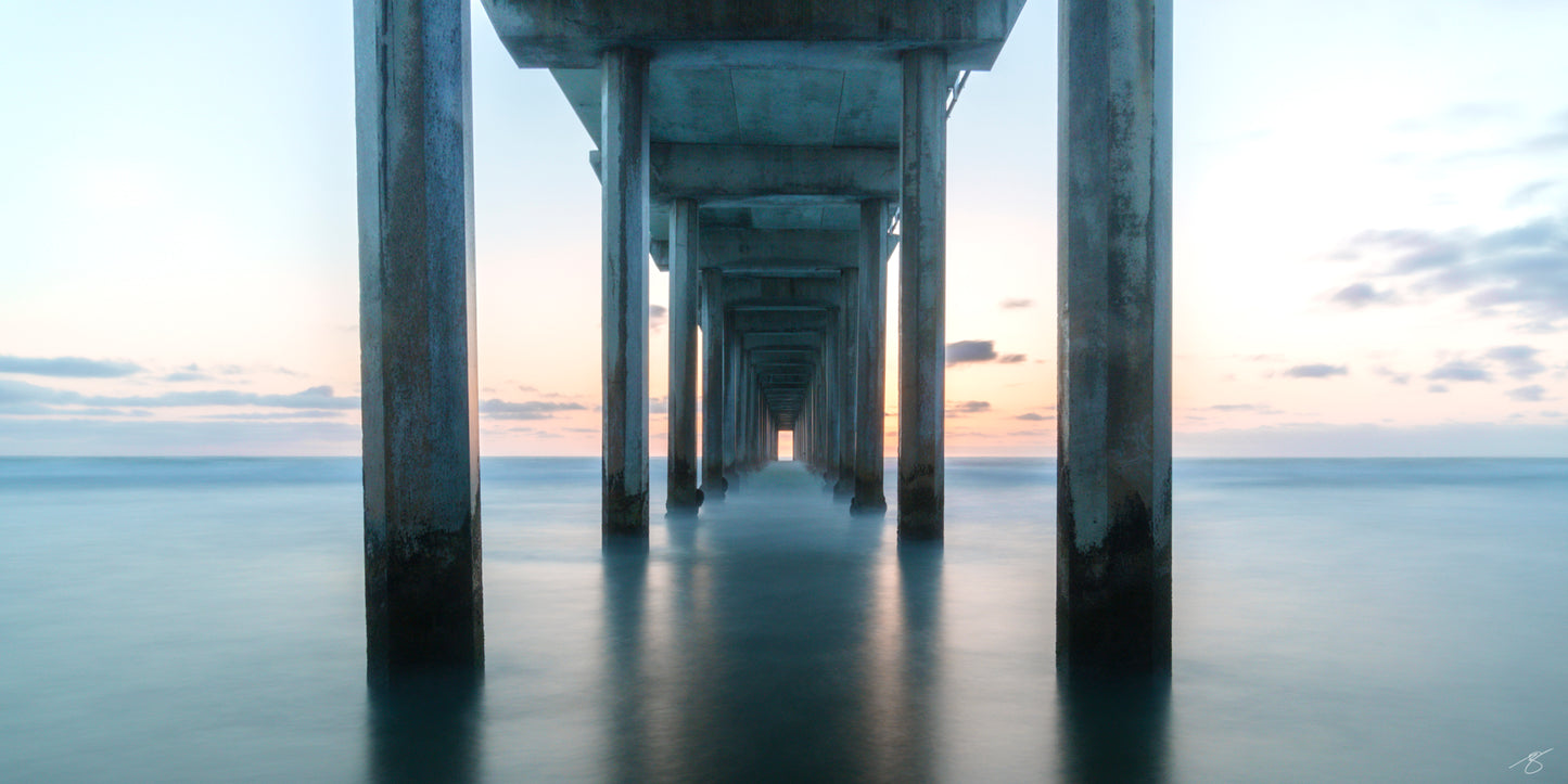 Long-exposure sunset beneath Scripps Pier featuring smooth, glassy ocean water, repeating concrete pillars, and a pastel coastal sky. A serene minimalist composition capturing calm light and perfect symmetry along the California coast.