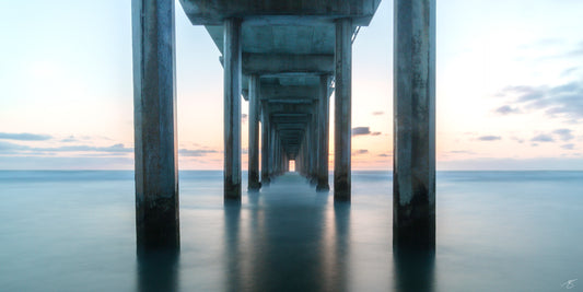 Long-exposure sunset beneath Scripps Pier featuring smooth, glassy ocean water, repeating concrete pillars, and a pastel coastal sky. A serene minimalist composition capturing calm light and perfect symmetry along the California coast.
