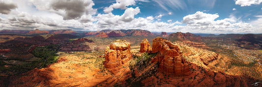 High-altitude panoramic photograph of Sedona featuring Cathedral Rock and surrounding red rock formations. Dramatic clouds cast shifting light across the desert landscape, revealing textures, canyons, and vibrant sandstone layers in a sweeping Southwest vista.
