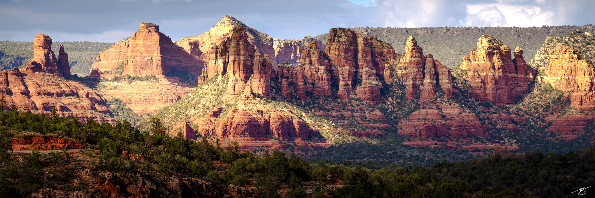 Wide panorama of Sedona’s iconic red rock cliffs and buttes illuminated by golden afternoon sunlight. Layers of sculpted sandstone rise above a forested desert foreground, creating a dramatic and detailed Southwest fine art landscape.
