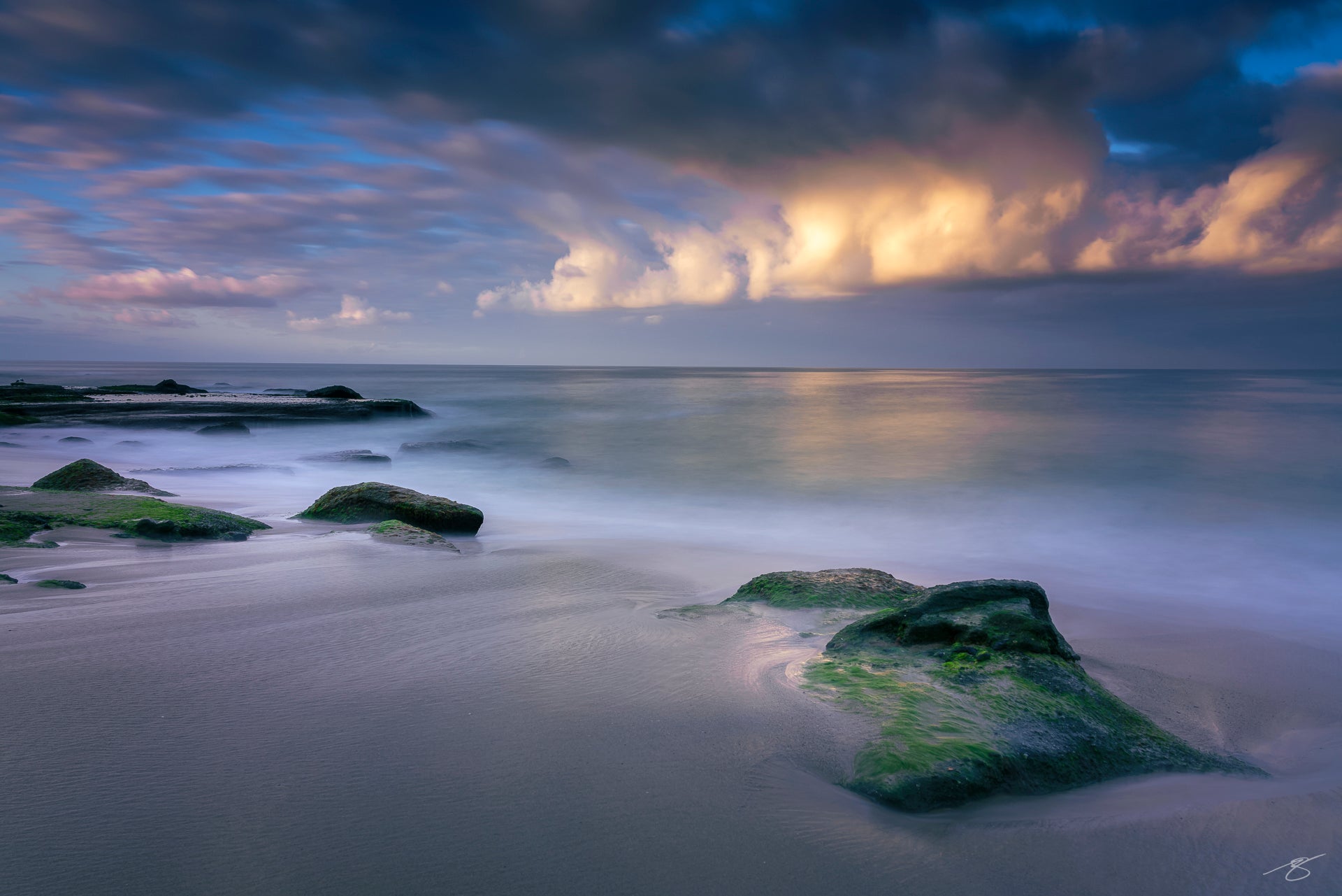 Long-exposure photo of a calm beach with mossy rocks, silky water, and glowing storm clouds at sunset over a tranquil ocean