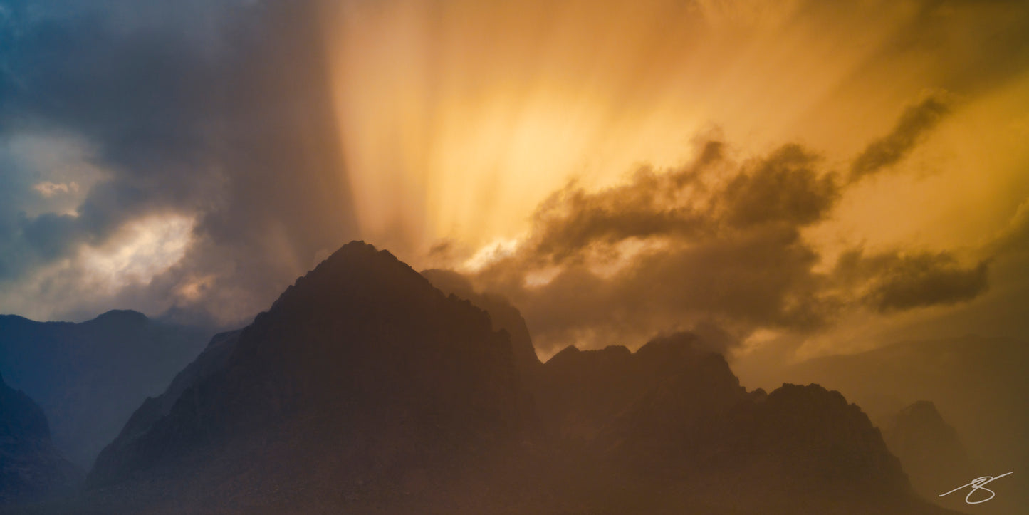 A dramatic desert landscape showing golden sunset light streaming through thick storm clouds above dark mountain silhouettes. The scene blends fiery orange tones with cool blue shadows, creating a powerful contrast of weather, light, and rugged terrain.
