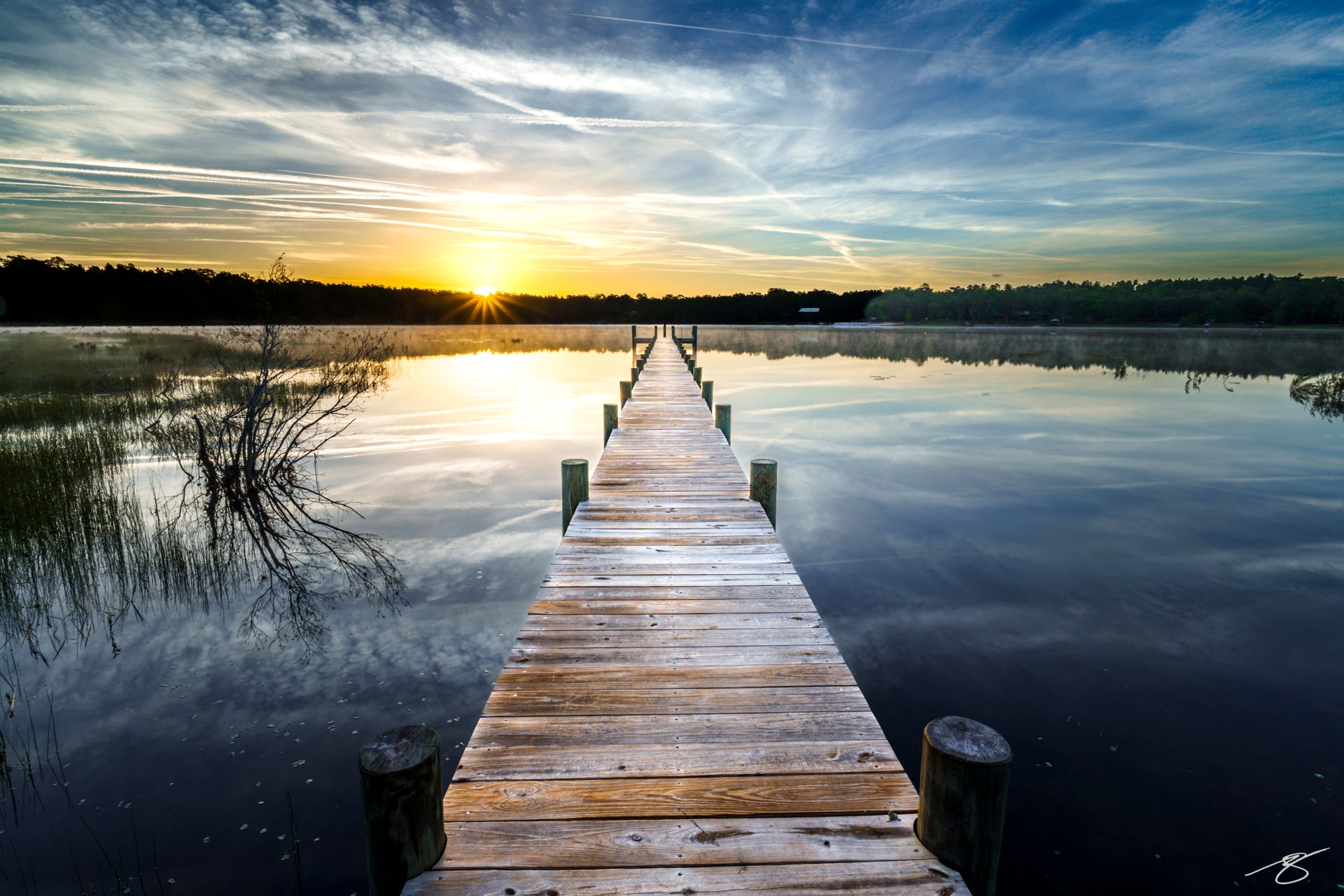 Wooden pier extending into a glassy lake at sunrise, golden sunstar on the horizon with soft clouds reflected on the water