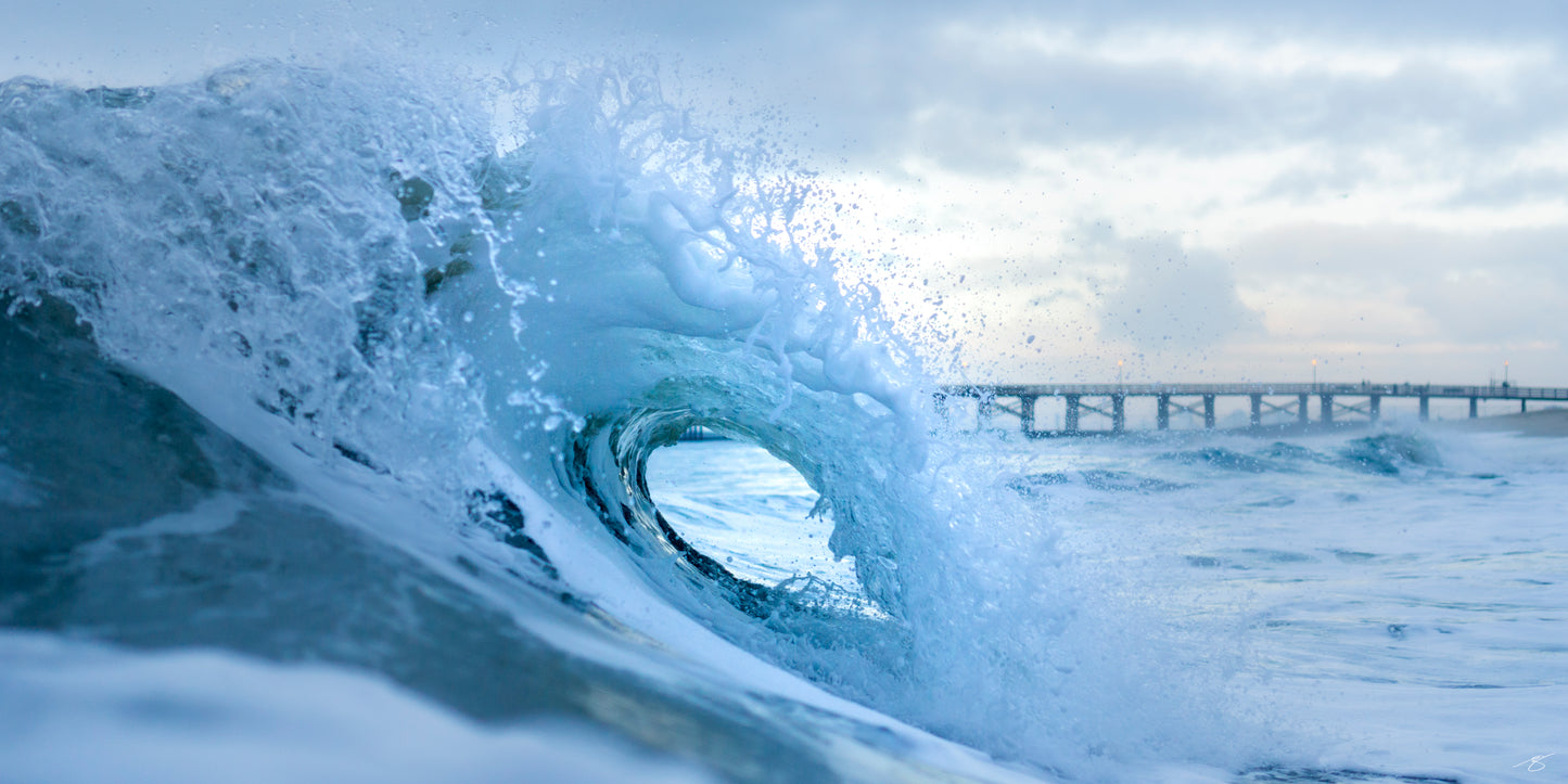 Powerful ocean wave curling into a translucent blue barrel at sunrise with a long pier in the distance. Soft morning light highlights the spray and motion, creating an energetic and atmospheric coastal surf photography scene.