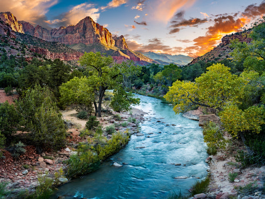Panoramic sunset of The Watchman above the Virgin River in Zion National Park, Utah, with glowing red cliffs, cottonwood trees, and a blue-green river winding through the canyon