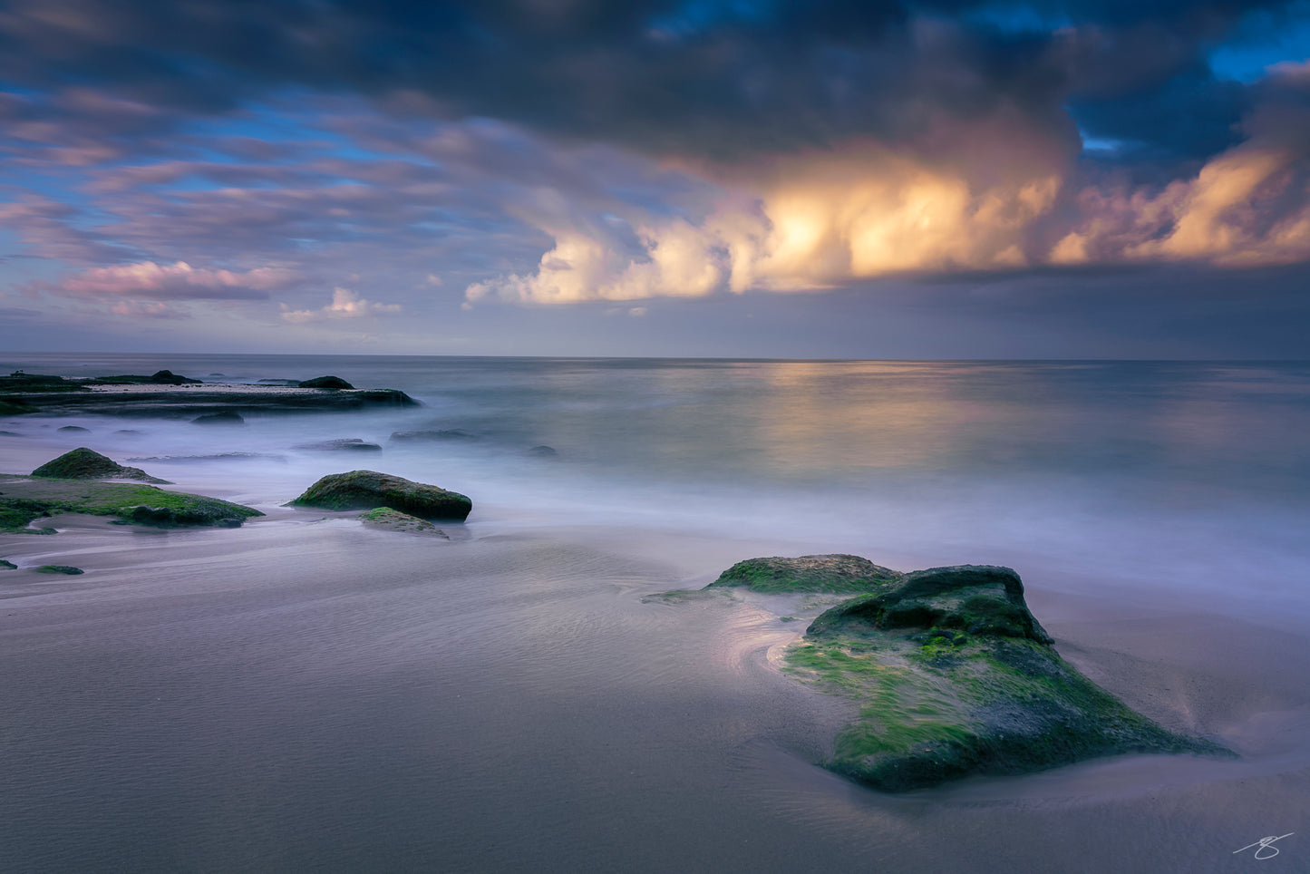 A minimalist coastal long-exposure photograph showing moss-covered rocks along a calm shoreline as smooth ocean water flows around them. Soft clouds glow above the horizon, creating a serene and atmospheric fine art seascape with muted blues and gentle highlights.