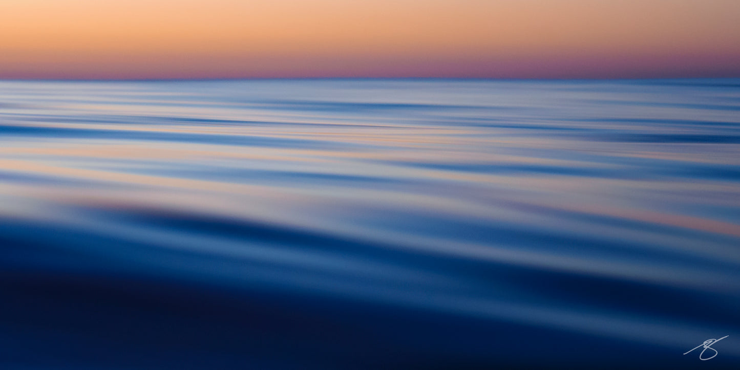 Abstract long-exposure photo of calm ocean surface with silky blue ripples beneath a peach and violet twilight sky; minimalist coastal wall art