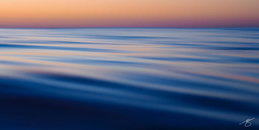 Abstract long-exposure photo of calm ocean surface with silky blue ripples beneath a peach and violet twilight sky; minimalist coastal wall art