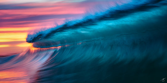 Abstract long-exposure photo of a teal ocean wave curling at sunset with magenta, pink, and golden sky streaks reflecting on the water