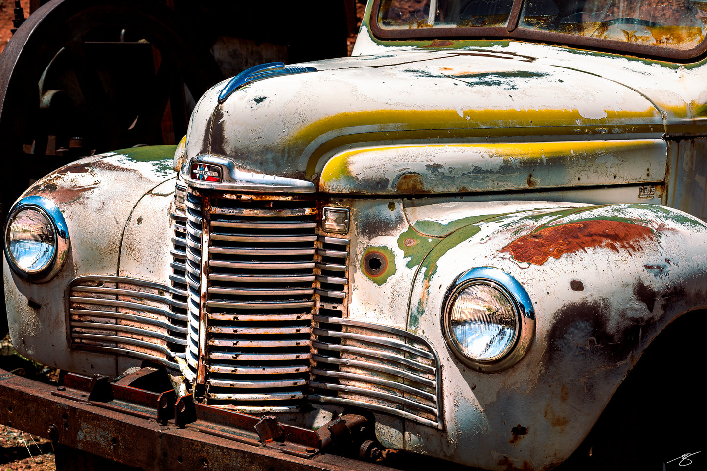 Close-up fine art photo of a vintage International Harvester truck featuring heavy patina, peeling paint in green and yellow tones, rust textures, chrome headlamps, and a classic grille illuminated by bright desert sunlight. A rustic piece of automotive history.
