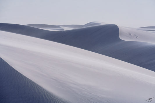Minimalist panoramic photo of smooth, wind-shaped sand dunes in soft blue and white tones under a pale sky; serene abstract desert landscape