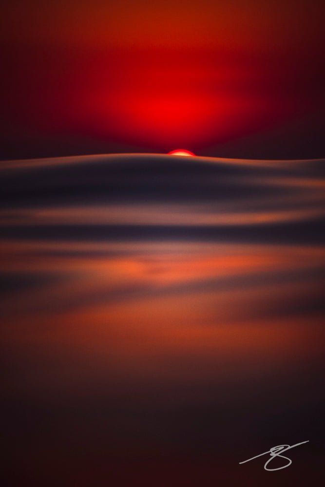 Minimalist seascape of a red sun dipping behind a smooth, blurred ocean horizon in deep indigo and crimson tones