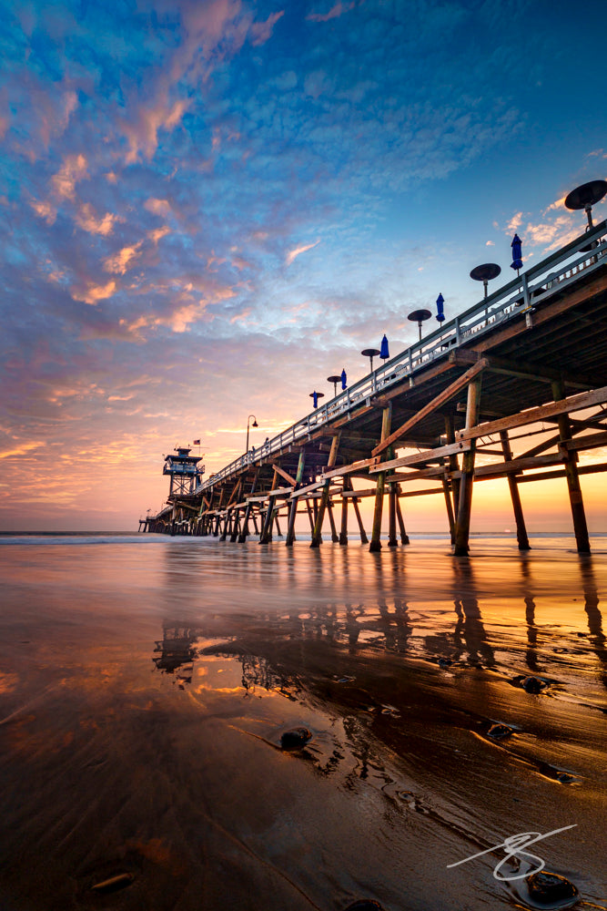 Sunset view of a wooden ocean pier with colorful clouds reflected on smooth, wet sand; serene coastal fine art photograph
