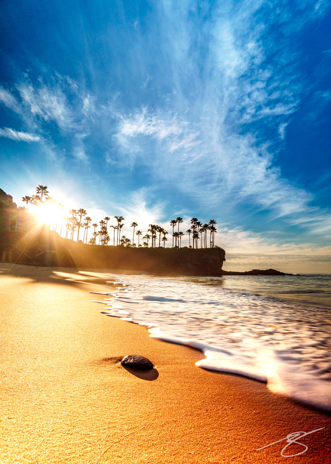 Sunrise over a palm-lined cove with golden sand, soft wave foam, and radiant sunbeams under a sweeping blue sky; coastal fine art beach photograph