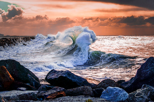 Dramatic wave exploding near a rocky jetty at sunset with backlit spray under storm clouds; coastal ocean fine art photograph