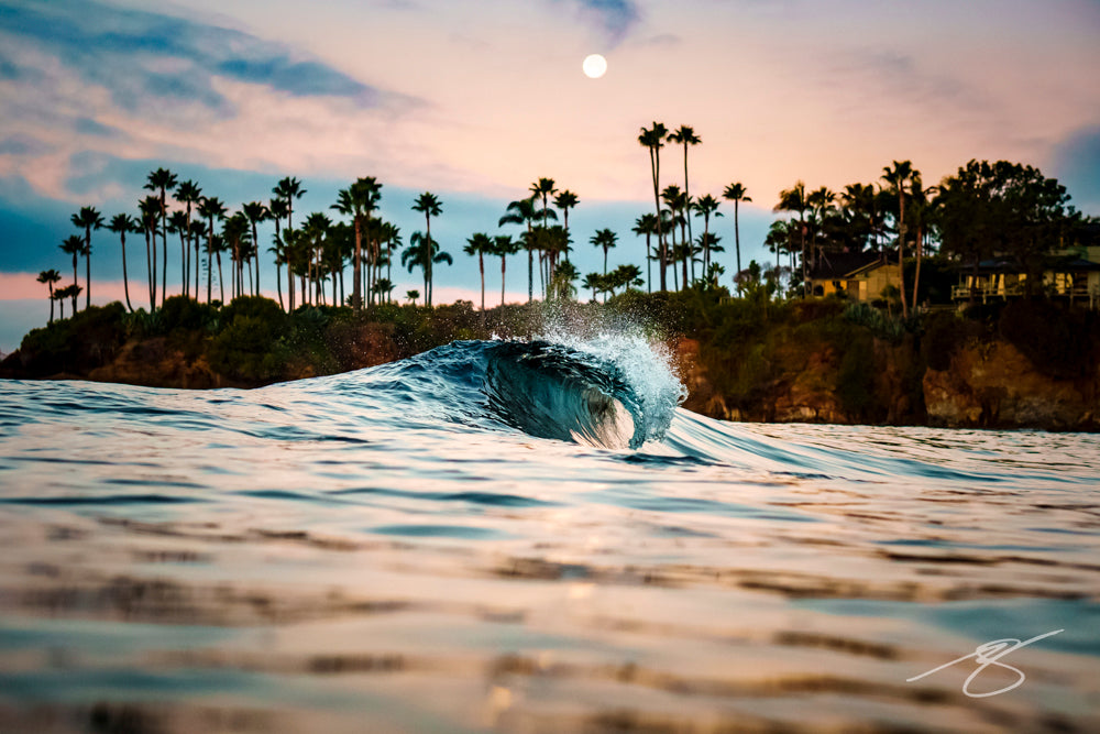 Waterline photo of a small barrel wave at dusk with a full moon above palm trees along a rocky cove; tranquil coastal fine art photograph