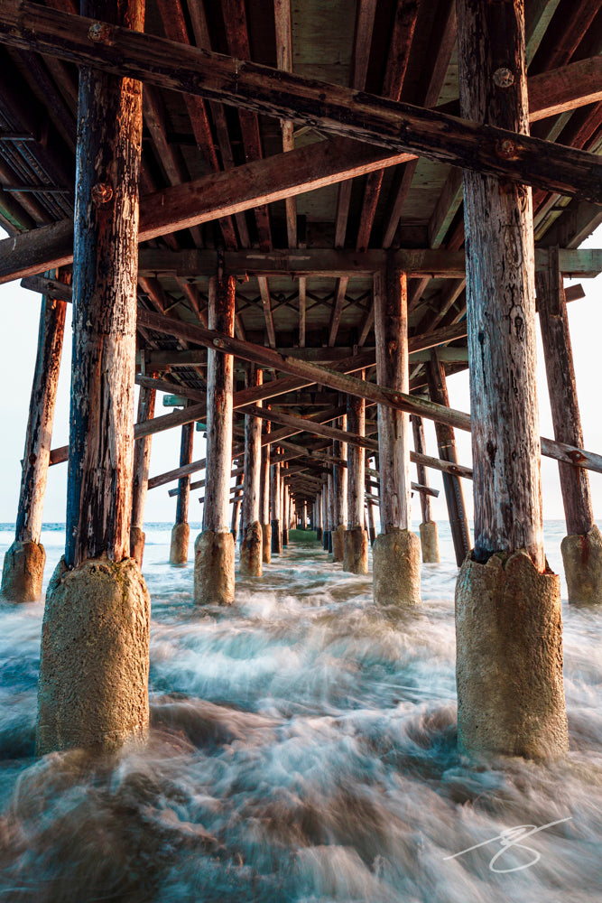 Long-exposure view beneath a wooden ocean pier with symmetrical pilings and blurred tidal water rushing through; dramatic coastal fine art photograph