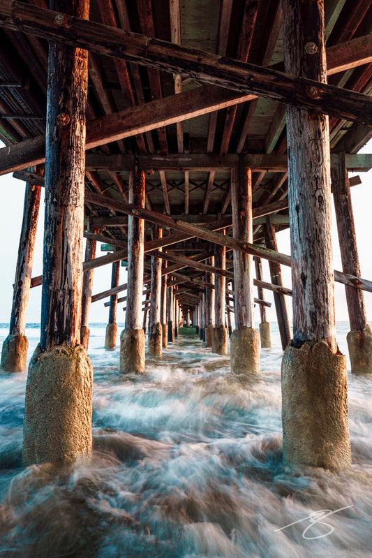 Long-exposure view beneath a wooden ocean pier with symmetrical pilings and blurred tidal water rushing through; dramatic coastal fine art photograph
