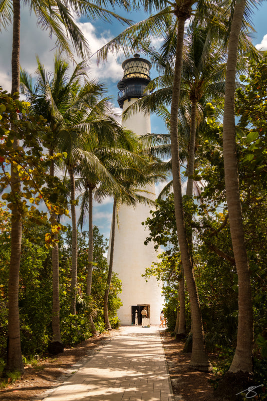 Palm-lined walkway leading to a white lighthouse under blue sky and soft clouds; tropical coastal fine art photograph, vertical orientation
